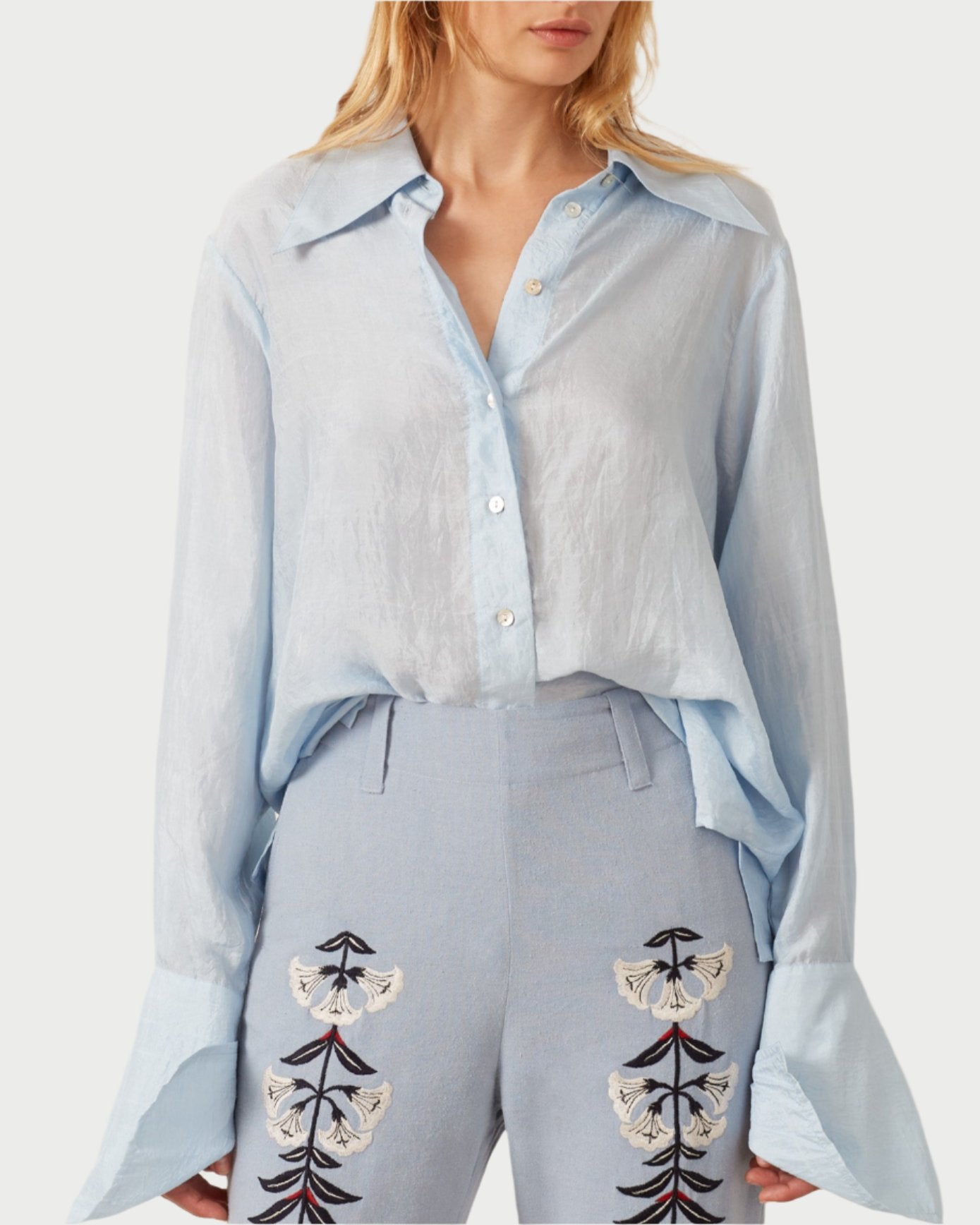 A person wears the Dolly Bluejay Silk Shirt, featuring an airy design in light blue with oversized cuffs, paired with pants inspired by Alix of Bohemia, embellished with floral embroidery. The ensemble exudes a casual yet stylish flair.