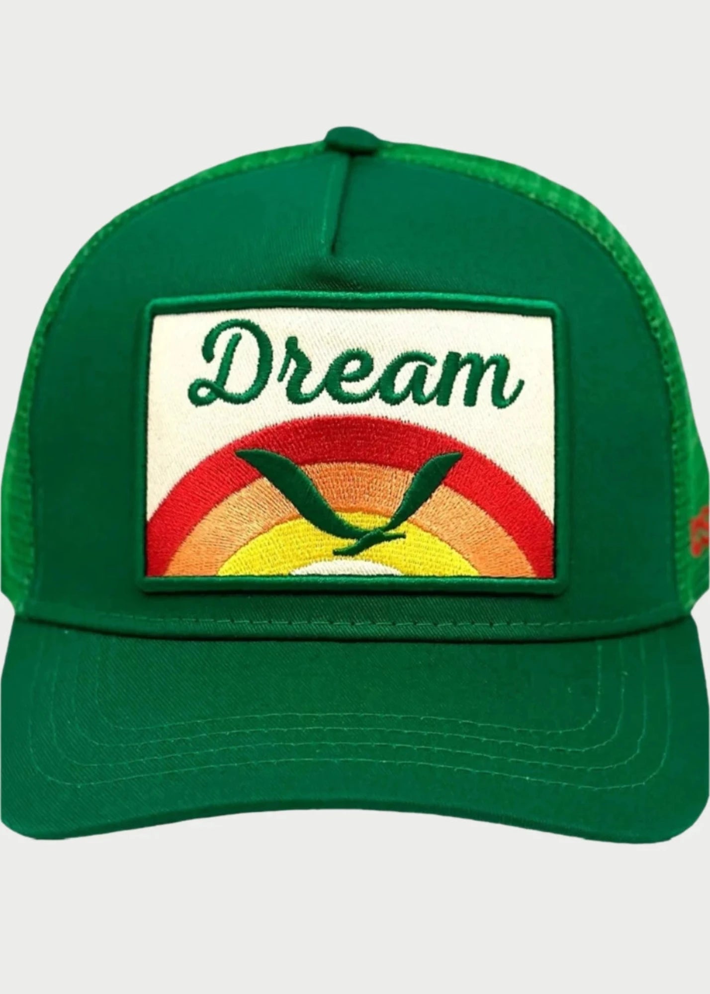 The Soulbyrd Dream Trucker Hat is an adjustable green cap featuring a mesh back and a front patch that reads "Dream," adorned with a stylized bird and a rainbow in shades of red, orange, and yellow.