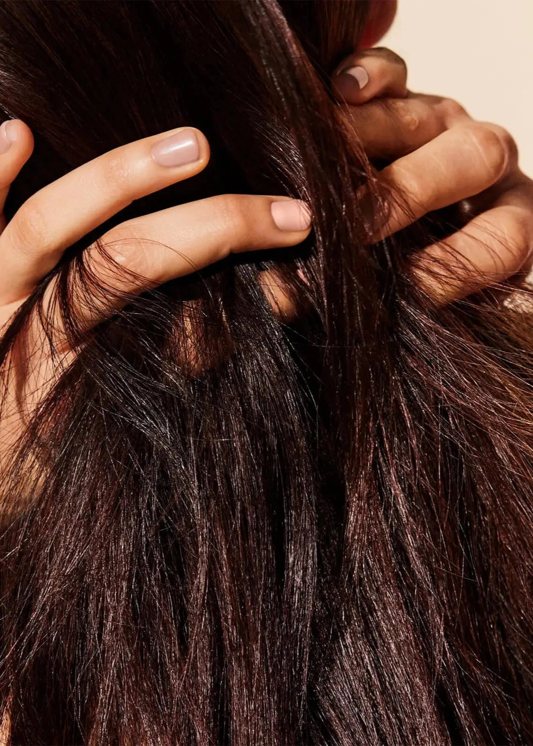 Close-up image of a person with long, dark brown hair, shown from behind. The person's hands, with natural nails, are placed on the back of their head, fingers interwoven through the hair treated with NUXE Huile Prodigieuse Riche dry oil. The background is a light beige color.