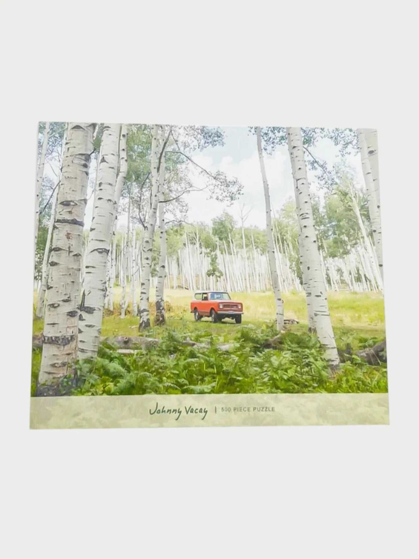 The "Johnny Vacay" 500 Piece Puzzles box showcases an image of a red SUV parked amidst a forest of white birch trees with vibrant green foliage. Ideal for family fun, this puzzle provides an engaging and enjoyable activity suitable for all ages.