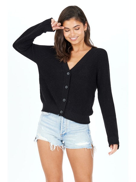 A person with dark hair is smiling and looking down while posing in a black oversized Raleigh Cardigan by One Grey Day, made of cotton fabric, paired with frayed light denim shorts against a plain white background.