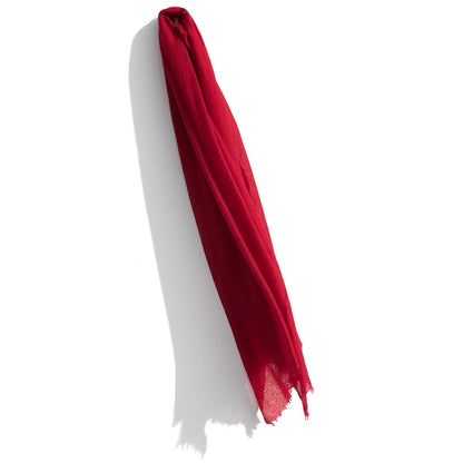 A vibrant red Whisperweight Cashmere Scarf by Meg Cohen hangs against a white background. The scarf's fabric appears lightweight and sheer, draping with soft folds and casting a delicate shadow on the wall. The bottom edge of the scarf is slightly frayed, showcasing its natural fibers.