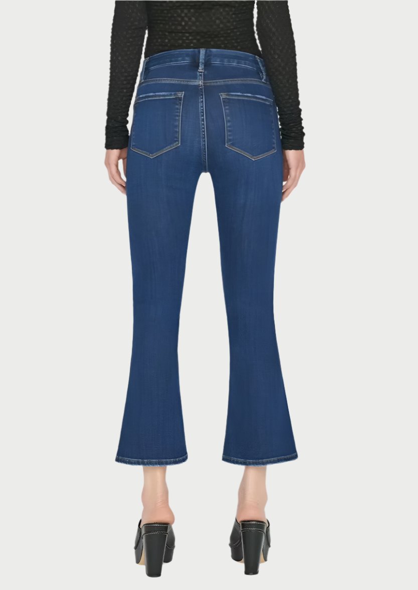 The person is seen from behind, wearing Frame's Le Crop Mini Boot Jeans in blue. These jeans are made from a sustainable stretch cotton blend and feature a contour mid-rise waist. They are paired with a black long-sleeve top, and the jeans showcase visible stitching and back pockets. The ensemble is completed with black heeled shoes against a plain background.