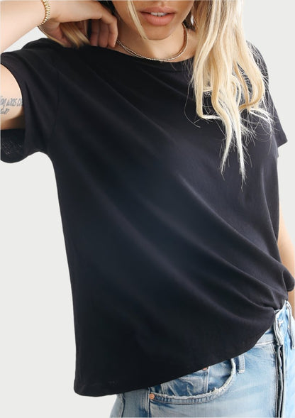 A person with long hair stands against a white background, wearing a perfectwhitetee Harley Boxy Crew in Multiple Colors and blue jeans. They touch the back of their neck with one hand, exposing a tattoo on their forearm.