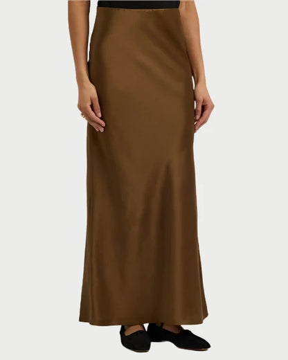 A person wearing the Silk Column Maxi Skirt by Frame, in a long brown shade, paired with black flat shoes. The skirt boasts a smooth, shiny texture and elegant column silhouette, reaching down to the ankles. The background is plain and light gray.