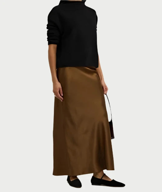 A person wearing a black long-sleeve top and the Frame Silk Column Maxi Skirt in brown, holding a small purse. They are also wearing black flat shoes against a plain white background.