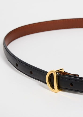 Close-up of the Petit Crescent Belt by Frame, crafted from black cowhide leather with a gold 'D'-shaped buckle. The inner side is brown, featuring visible punch holes for adjustment. The belt is elegantly displayed on a white surface, showcasing its gold-tone hardware.