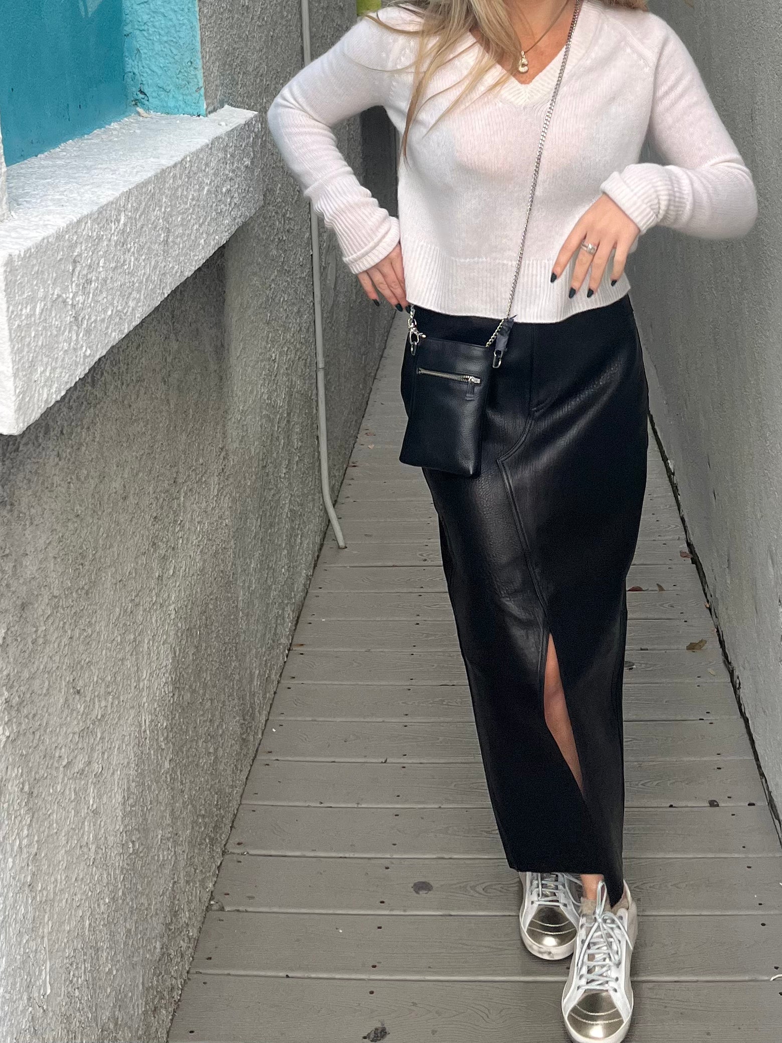 A person with long hair is walking down an alley. They are wearing a white long-sleeve shirt, a black Long Leather Skirt from Frame with a front slit in lamb leather, silver sneakers, and a small black crossbody bag. The background features a wall with blue and white paint.