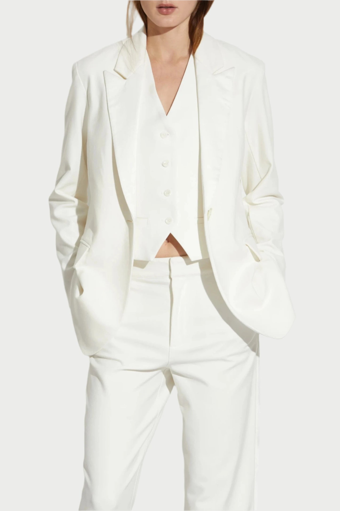 A person dressed in the elegant Saint Art NY Dylan Tuxedo Jacket in Ivory poses against a plain white background, wearing matching ivory pants and shoes. With their hands in their pockets and long hair flowing, they exude sophistication appropriate for formalwear.