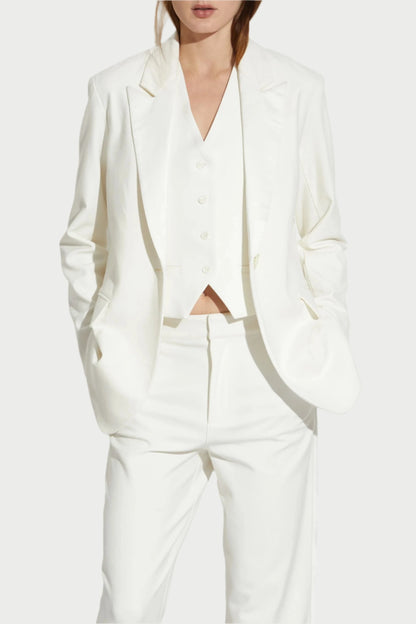 A person dressed in the elegant Saint Art NY Dylan Tuxedo Jacket in Ivory poses against a plain white background, wearing matching ivory pants and shoes. With their hands in their pockets and long hair flowing, they exude sophistication appropriate for formalwear.