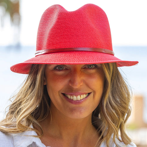 A person with long, wavy hair is smiling at the camera. They are wearing a red Summer Hat from Travaux en Cours, featuring a thin leather band and a white top. The background is slightly blurred, showcasing a sunny outdoor scene with blue sky and hints of greenery and water.