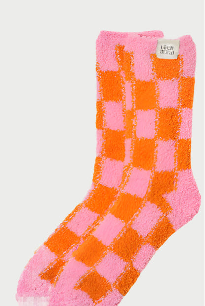 LOCAL BEACH's Cozy Socks showcase a fluffy, pink, and orange checkered pattern with a small label on top. These feather knit gems are perfect for adding warmth to your day when laid flat against a plain background.