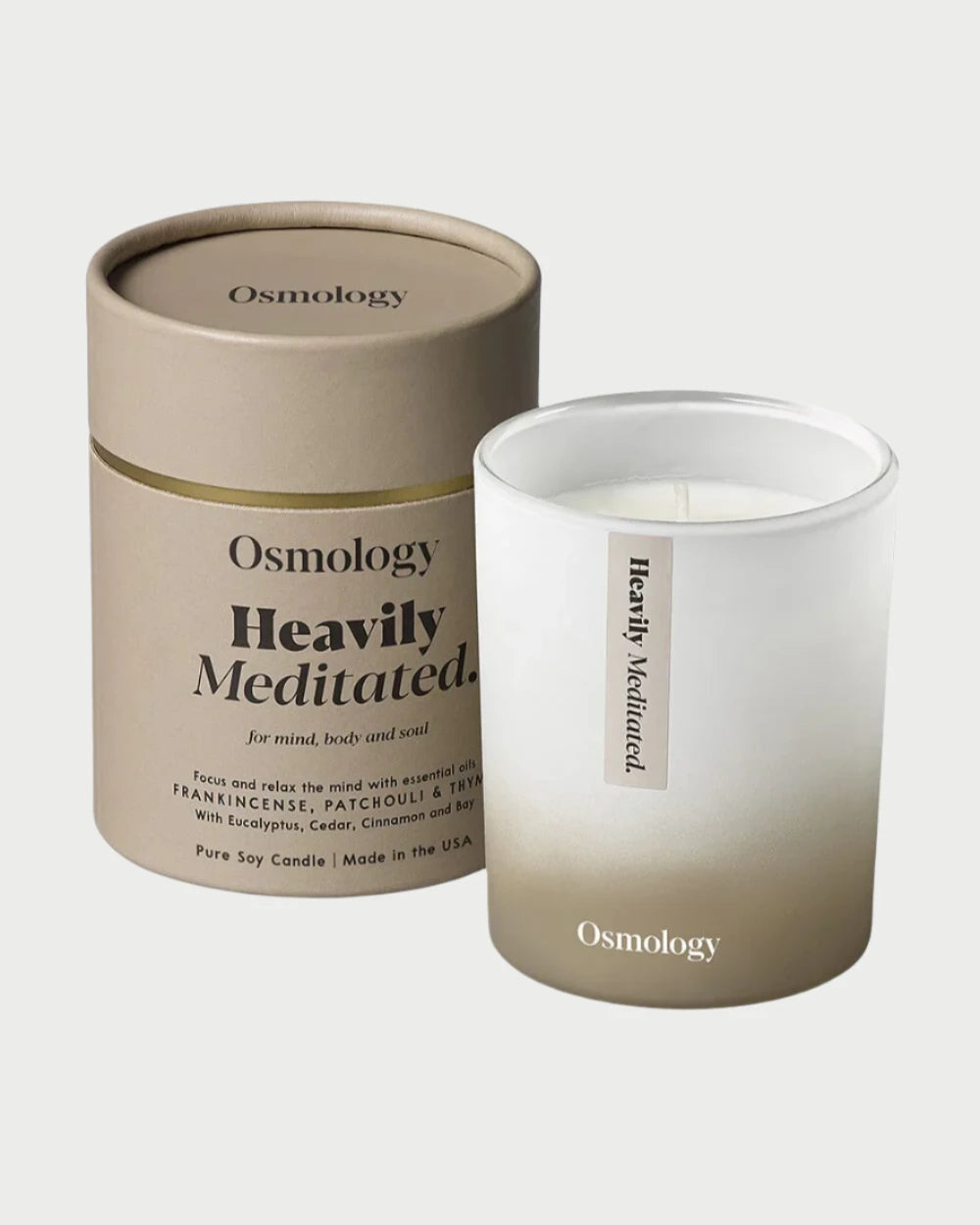 The Osmology Candle is presented with its cylindrical brown and beige packaging. This white candle, branded by Osmology, is encased in a gradient glass container. Crafted from pure soy and essential oils, it features notes of patchouli to enhance your aromatherapy experience.