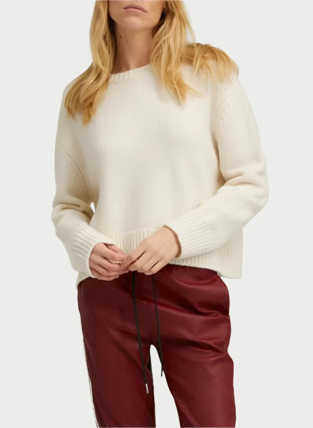 A person dressed in an Ivory Heavy Crewneck Sweater and SPRWMN burgundy leather pants stands against a plain background. Their long, wavy blonde hair cascades around them, and their hands are gently clasped in front, exuding effortless elegance.