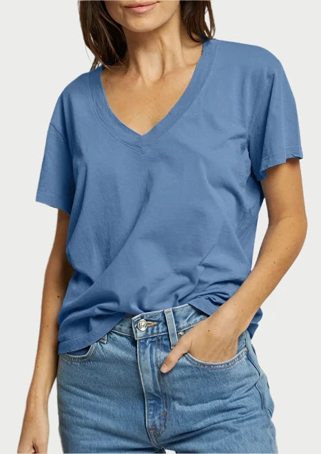 A woman with shoulder-length brown hair wearing the Hendrix Tee in Carolina Blue by perfectwhitetee and light-wash jeans stands against a plain light gray background. She has a neutral expression, and her hands are resting casually in her jean pockets.