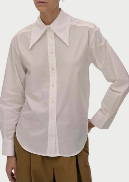 A person with their hair tied back is wearing a 100% cotton Dara Shirt from Herskind with an exaggerated collar and brown trousers. They are standing against a plain background with one hand resting on their hip, looking in the distance with a neutral expression.