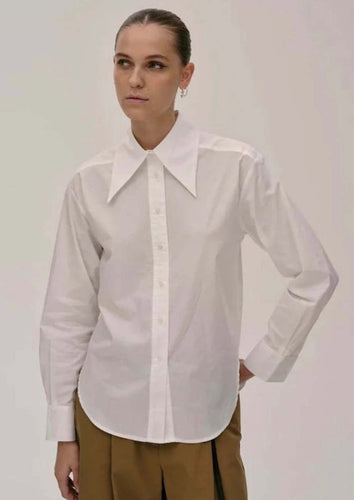 A person with their hair tied back is wearing a 100% cotton Dara Shirt from Herskind with an exaggerated collar and brown trousers. They are standing against a plain background with one hand resting on their hip, looking in the distance with a neutral expression.