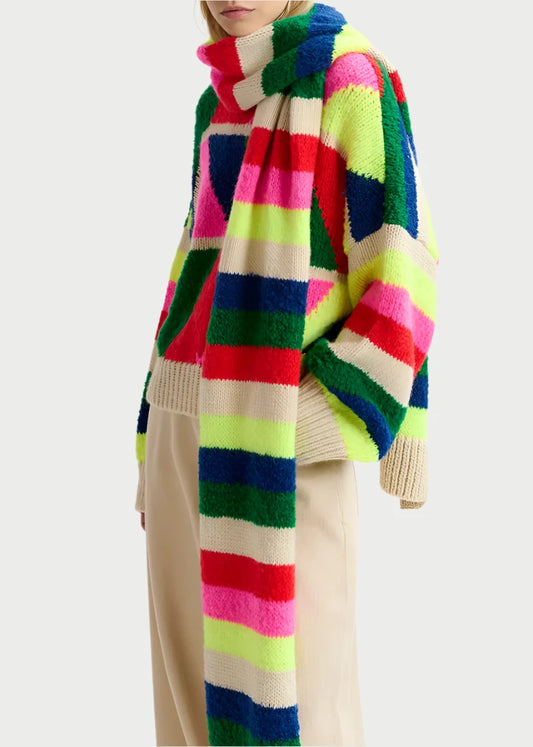 A person with blonde hair is wearing a colorful, striped sweater featuring intricate intarsia-knit patterns, paired with the Garis Knitted Scarf from Essentiel Antwerp in vibrant hues of green, pink, blue, and yellow. They are also wearing beige pants against a plain white background.