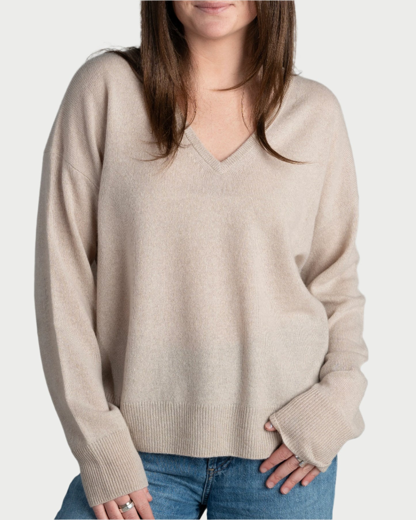 A person wearing the Archer Boyfriend Cashmere V-Neck sweater in beige and blue jeans stands with one hand in a pocket. The background is plain and neutral, with their long brown hair partially covering their shoulders.