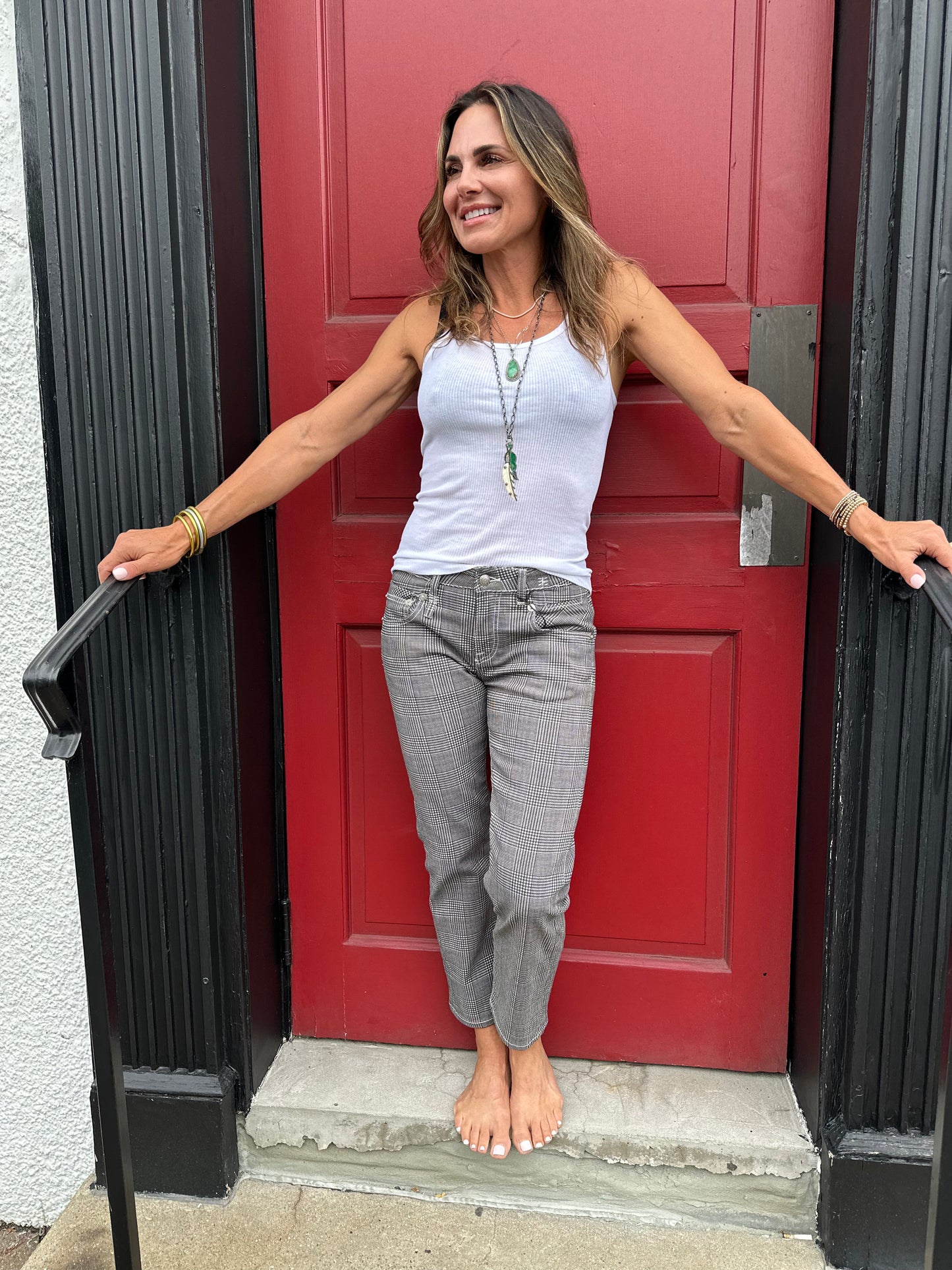 A woman stands barefoot on a doorstep in front of a red door. She is wearing a white, 100% cotton Ribbed Tank from RE/DONE, gray plaid pants, and multiple bracelets. Her long hair is down, and she is smiling with her arms resting on the door frame.