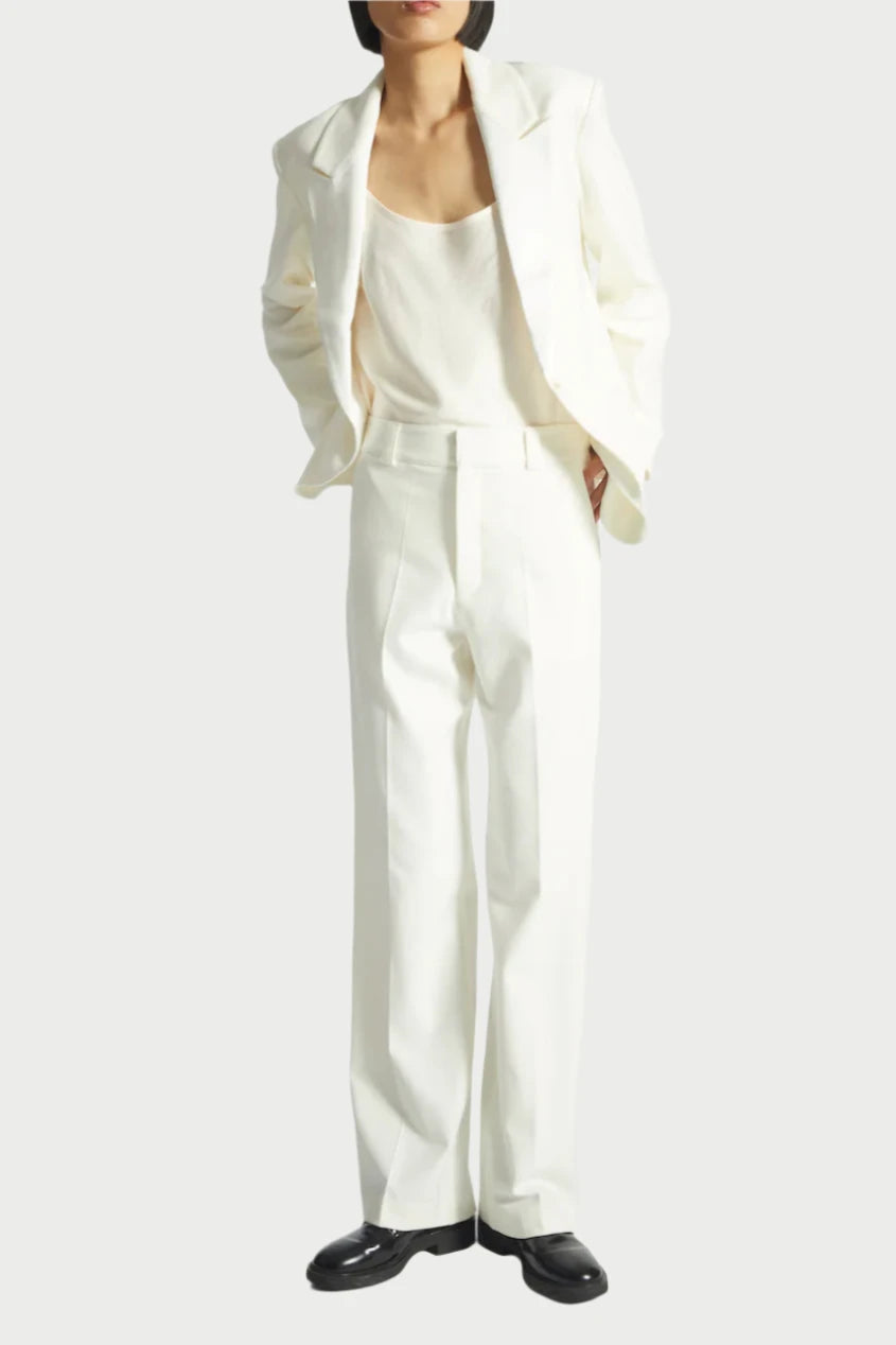 A person with short hair poses confidently against a plain background, wearing the Dylan Tuxedo Jacket in Ivory by Saint Art NY. The white suit, featuring a charmeuse satin lapel, is paired with a tank top and black shoes, as they tuck their hands into the jacket pockets.