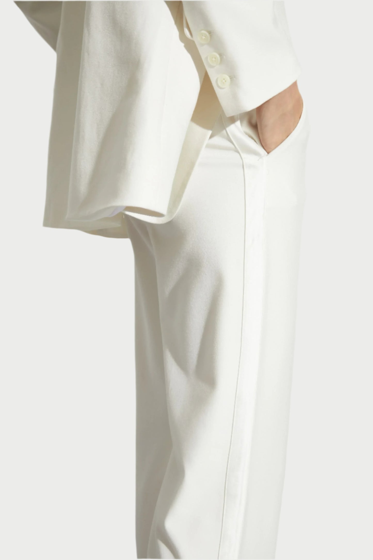 A person clad in a white suit made from breathable fabric stands with their hand in the pocket. The Cait Low-Rise Tuxedo Pants by Saint Art NY drape stylishly, and the image skillfully highlights the essence of tuxedo dressing, emphasizing the midsection and lower half of the ensemble.