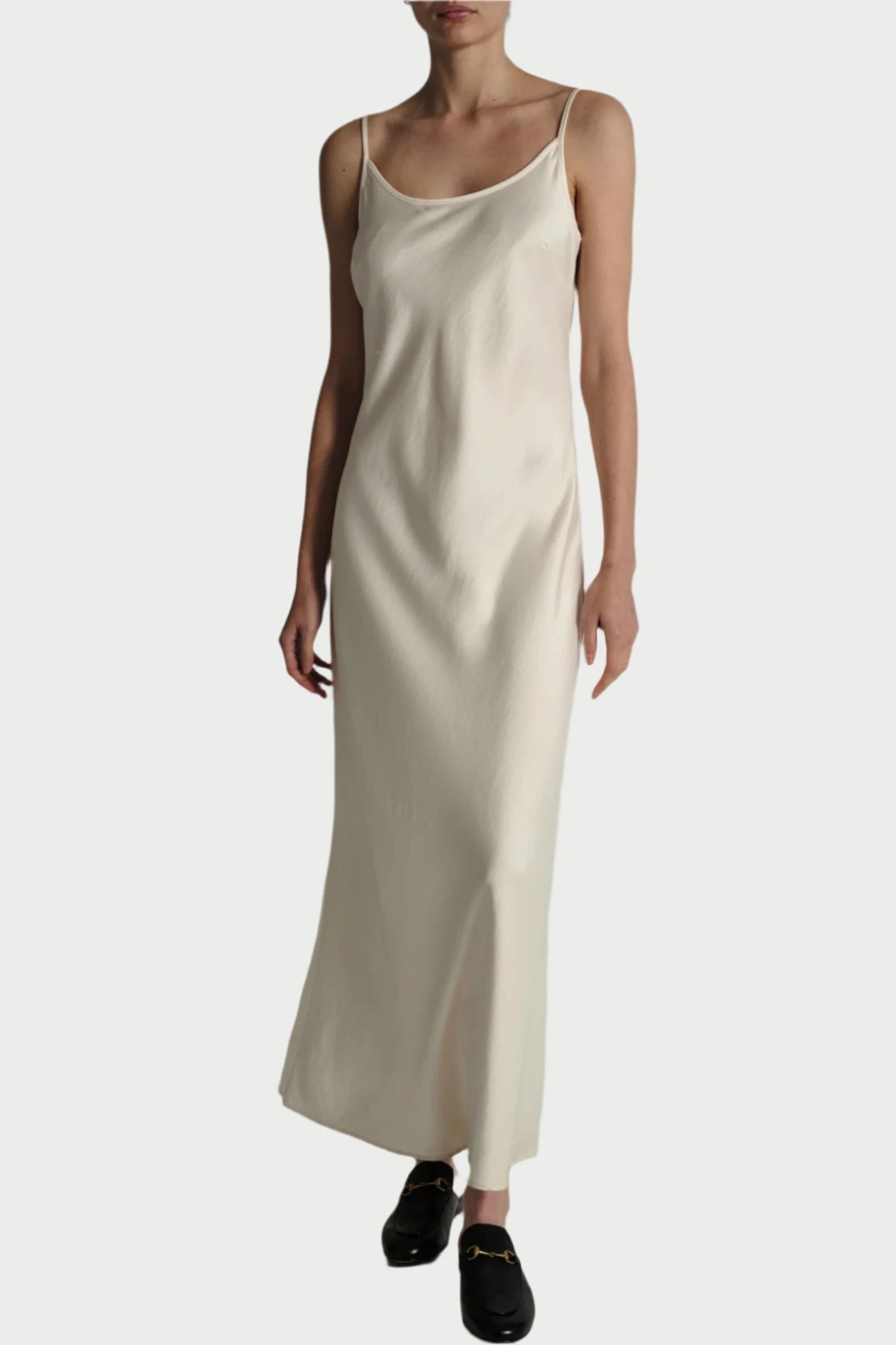 A person is wearing a long, sleeveless Saint Art NY Ivory Haley Slip Dress with spaghetti straps and black loafers. The dress has a simple, elegant design and appears to be made of silky, smooth, heavyweight charmeuse satin that is wrinkle-resistant. The background is plain and light-colored.