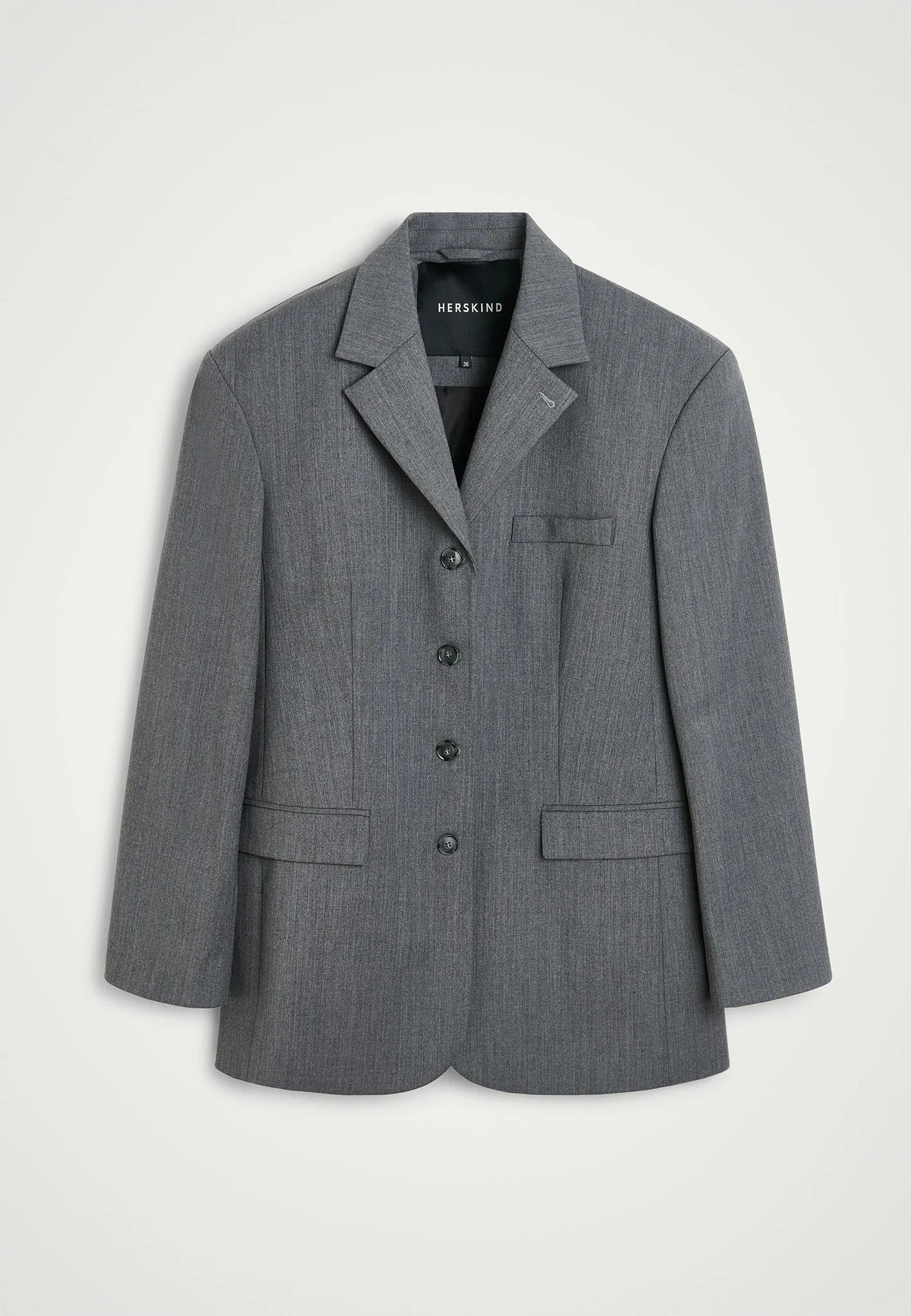 A gray, single-breasted Yves Blazer by Herskind hangs elegantly against a white background. This blazer, inspired by men's suiting, features a notched lapel, three buttons down the front, a breast pocket on the left side, and two flap pockets at hip level. The label inside the collar reads "HERSKIND.