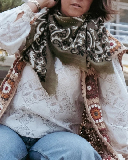 A person with shoulder-length dark hair is wearing sunglasses, a white lace blouse, a colorful crocheted vest, and blue jeans. They accessorize with a Maison Lecomte Flament Wool Cashmere Bandana Scarf, adding flair to their ensemble. The background showcases a building with a yellow-striped awning and lush greenery.