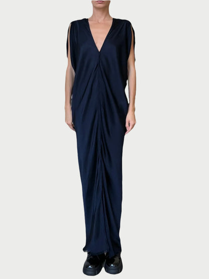 A person stands against a plain background wearing the Leah Dress by Jaga LA, a long, dark blue draped dress made from high-quality fabric. The deep V-neckline adds elegance, while the black shoes perfectly complement the look. This versatile piece is perfect for any occasion.