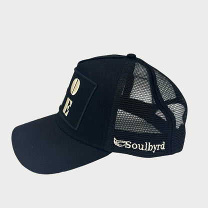 A black mesh trucker cap with a solid front panel displaying the word "LOVE" in bold letters and featuring "Soulbyrd" in white lettering on the side.