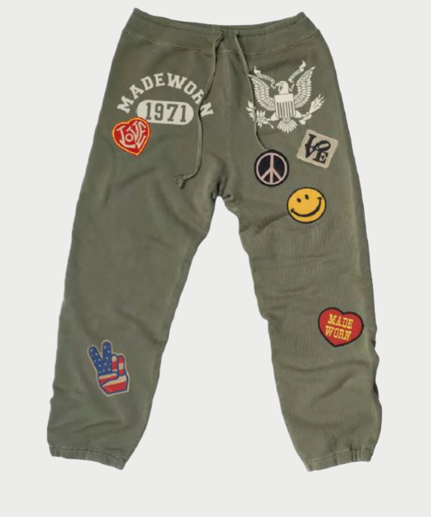 The MadeWorn 1971 Sweatpants by Madeworn are olive green and adorned with sewn patches like an eagle, peace sign, smiley face, and heart. The text includes "Madeworn" and "1971." These pants are vintage-inspired with distressed details on 100% cotton fabric.