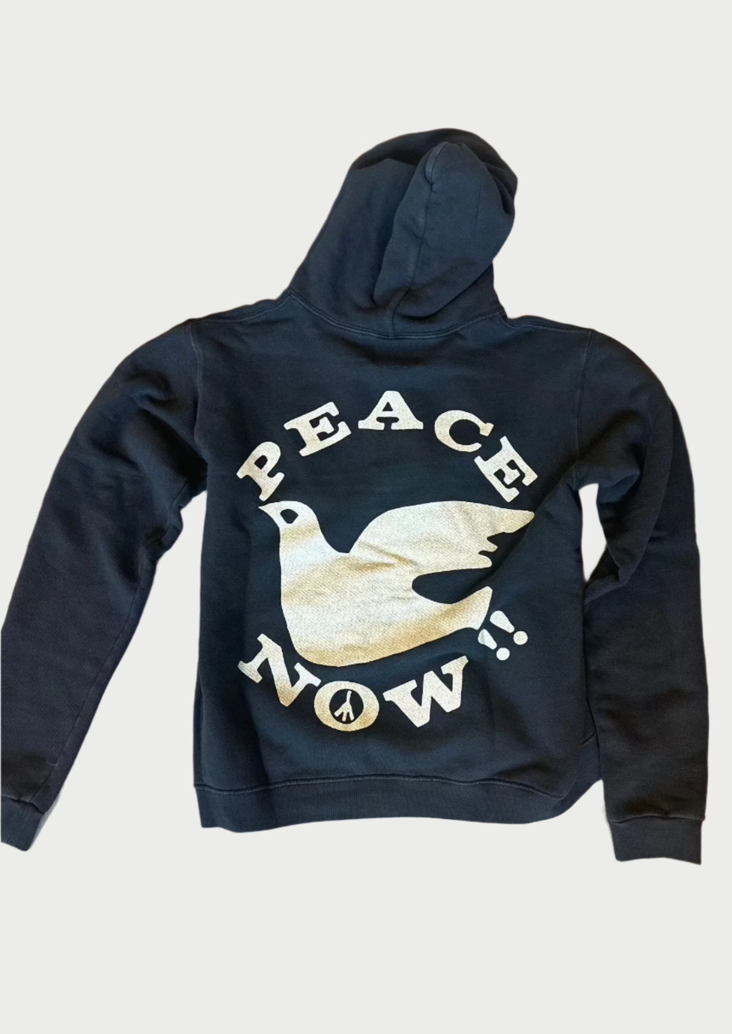 A vintage-inspired design, this black Peace Now Hoodie by Madeworn with a hood lying flat on a wooden floor showcases a large graphic of a white dove and the text "PEACE NOW!" in bold letters on the back, with a small peace symbol below near the dove's tail.