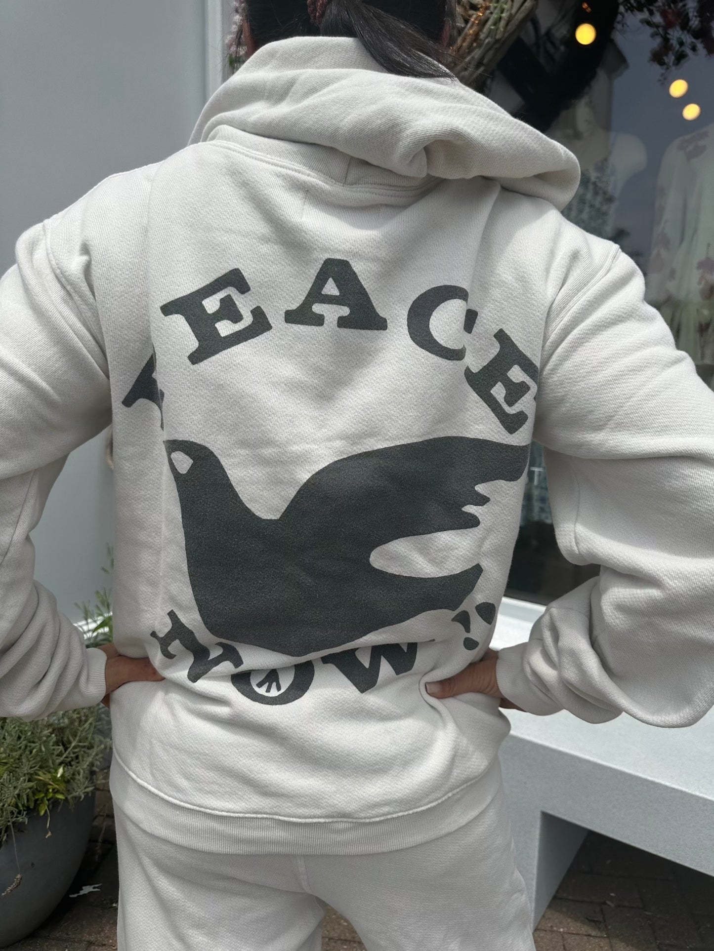 A person wearing a light gray Peace Now Hoodie by Madeworn with the words "PEACE NOW" and a black dove printed on the back is standing outdoors. The person is facing away from the camera. The background shows part of a building and some greenery, adding to the vintage inspired design.