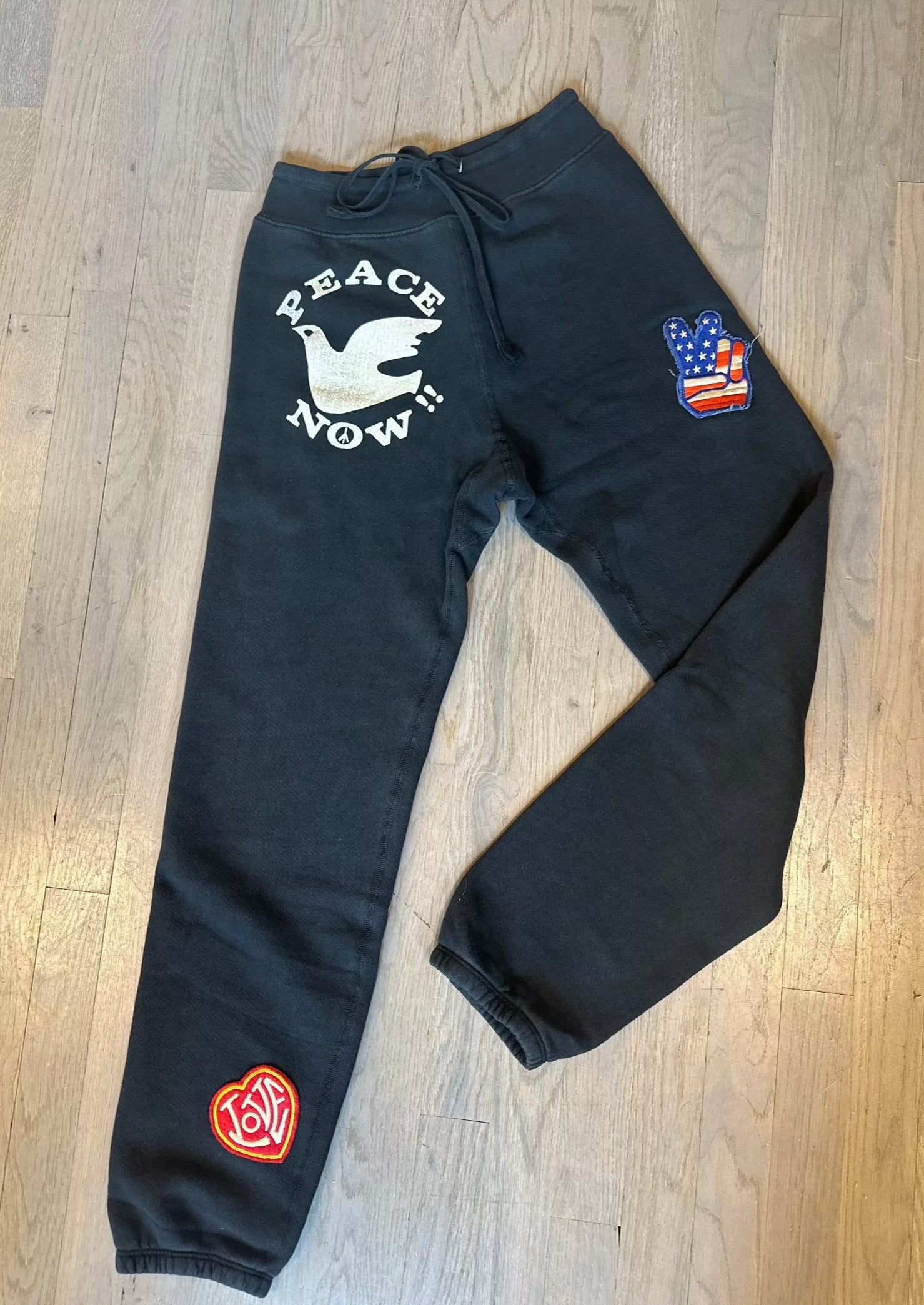 A pair of handmade Madeworn Peace Now Sweatpants is displayed on a wooden floor. The left leg features a white dove and the text "Peace Now!" on the thigh, and a circular red logo near the ankle. The right leg has a patch with an American flag and a peace sign on the thigh, all in a vintage unisex fit.