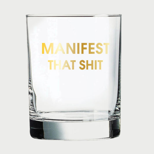 A Chez Gagné More Rocks Glass features gold foil lettering reading "MANIFEST THAT SHIT" on a clear glass, elegantly set against a plain white background.