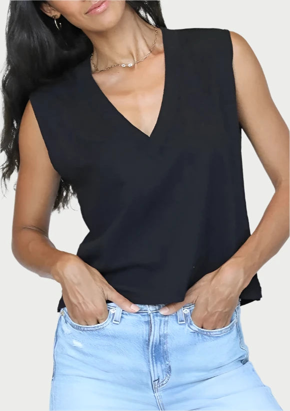 A person in a black Margot V Neck Sleeveless Tee by perfectwhitetee and light blue jeans, with hands casually in pockets. Their long, wavy dark hair complements the top, and they accessorize with a delicate necklace and hoop earrings, standing against a plain background.