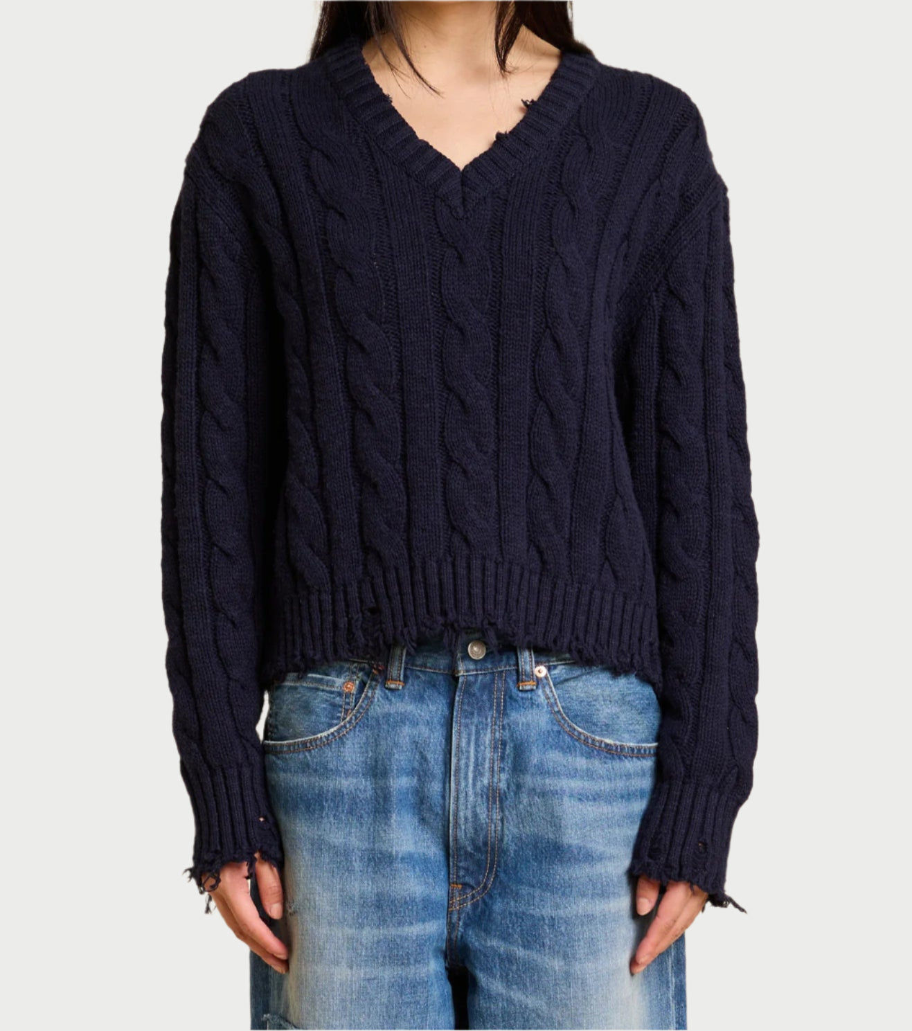 A person in a dark blue Denimist Cable V-Neck Sweater and blue jeans stands against a plain background. This timeless piece features ribbed cuffs, hem, and subtle distressed cable detailing. The person isn't fully visible in the image.