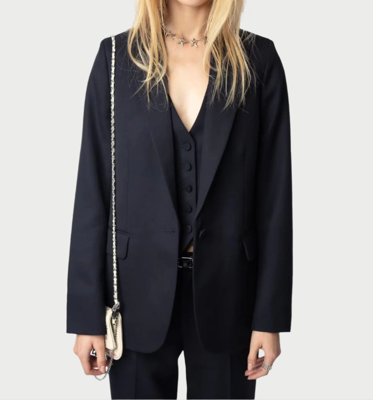 A person with long blonde hair is styled in a Zadig & Voltaire Tailored Navy Blazer over a black suit with a vest and star necklace, complemented by a chain-strap bag against a plain, light-colored background.