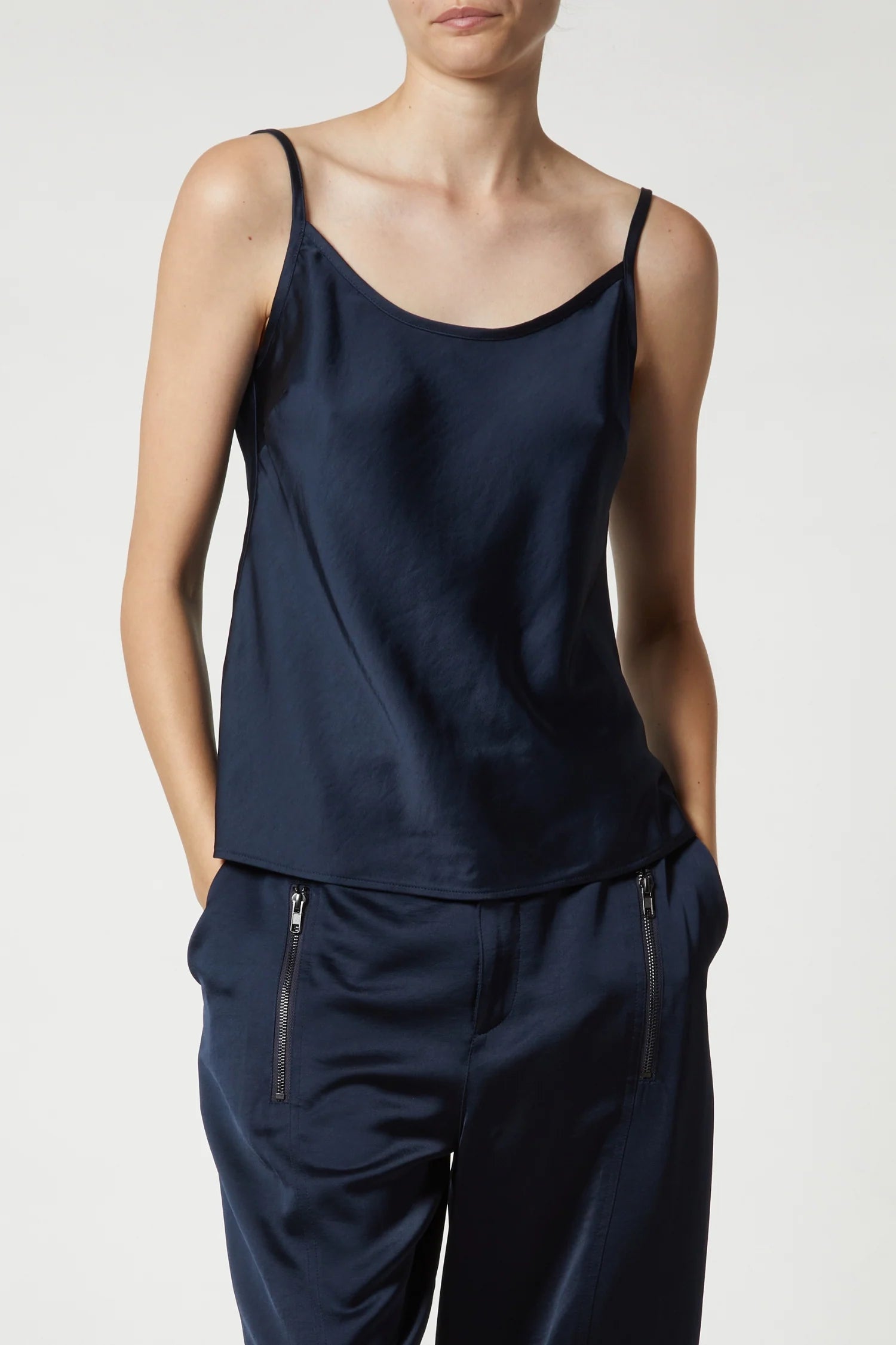 A person in a navy Charmeuse Camisole by Saint Art NY with matching trousers featuring zippered pockets stands against a plain background, hands in pockets, adding versatile color to their look. Their face is partially visible.