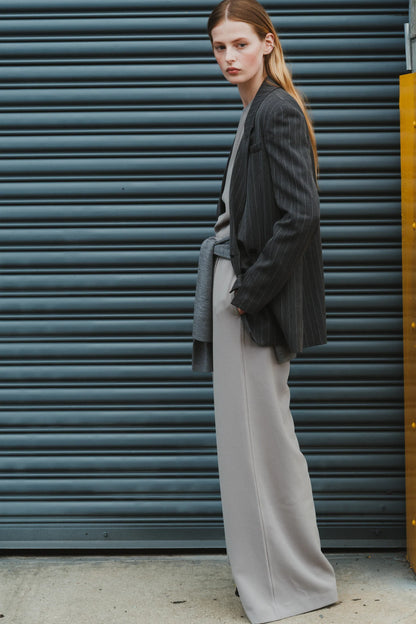 A person with long hair stands against a blue industrial metal door, wearing a black striped blazer over a grey coat and Saint Art NY's Neve Crepe Pant in Powder Grey. Showcasing relaxed tailoring, they have one hand in their pocket and are looking intently at the camera.