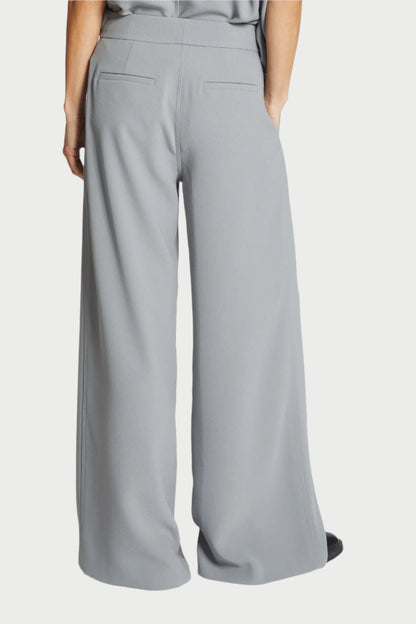 A person standing with their back to the camera wears Saint Art NY's Neve Crepe Pant in Powder Grey, high-waisted with wide legs and featuring relaxed tailoring, including two back pockets without visible buttons. The trousers are paired with a matching light grey top tucked in—a timeless essential for any wardrobe.