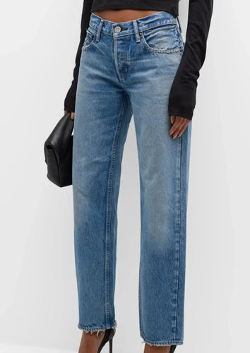 A person wearing high-waisted, light-wash Cheval Straight-Low Jean from Moussy with frayed hems, a black long-sleeve top, and black high-heeled shoes. They are holding a small black pouch in their left hand. The background is plain white.