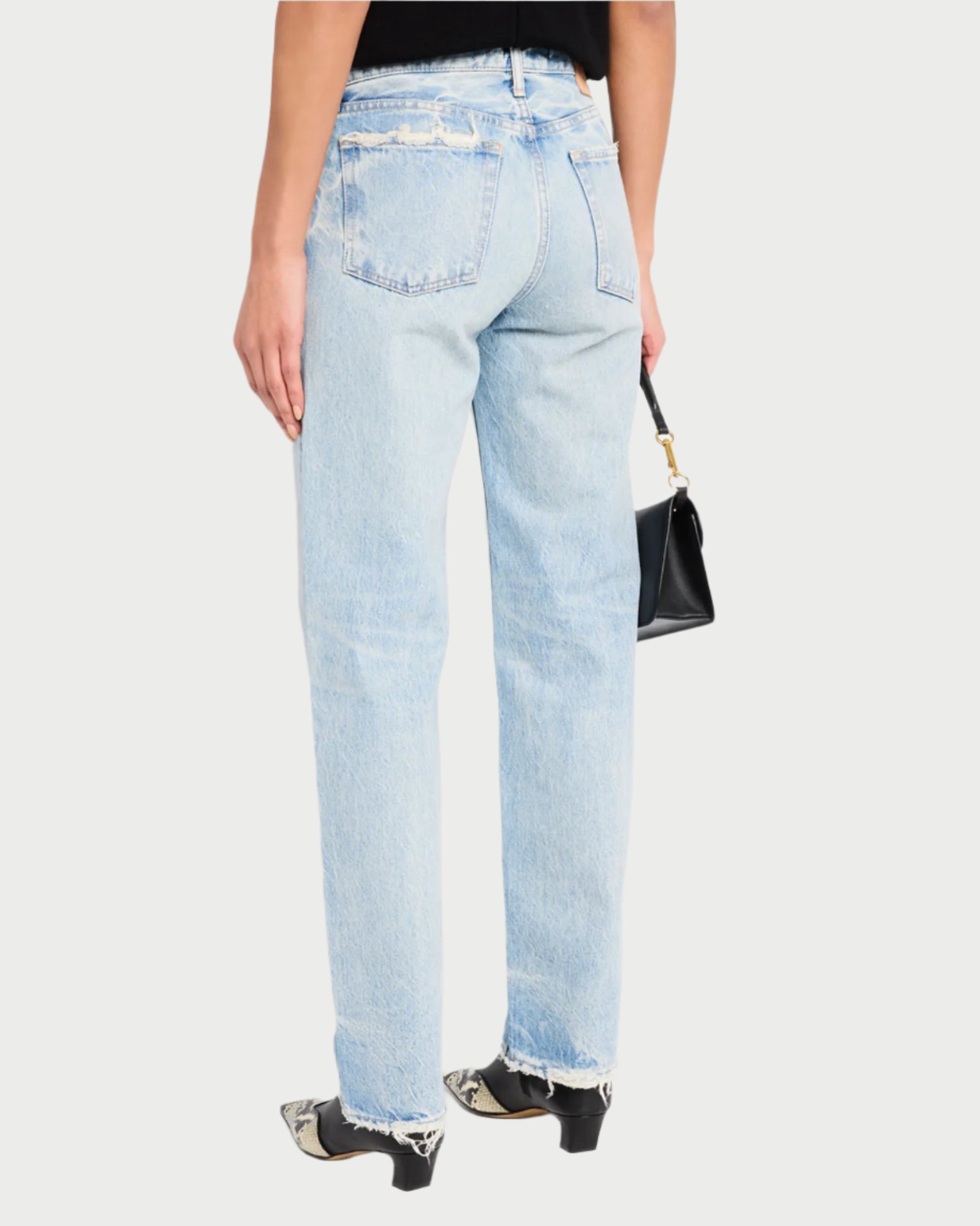 A person is wearing Moussy's Wildwood Straight Leg Jeans in faded light-wash with frayed hems, a black top, and snakeskin-patterned pointed shoes. They're holding a small black handbag and stand against a plain white background.