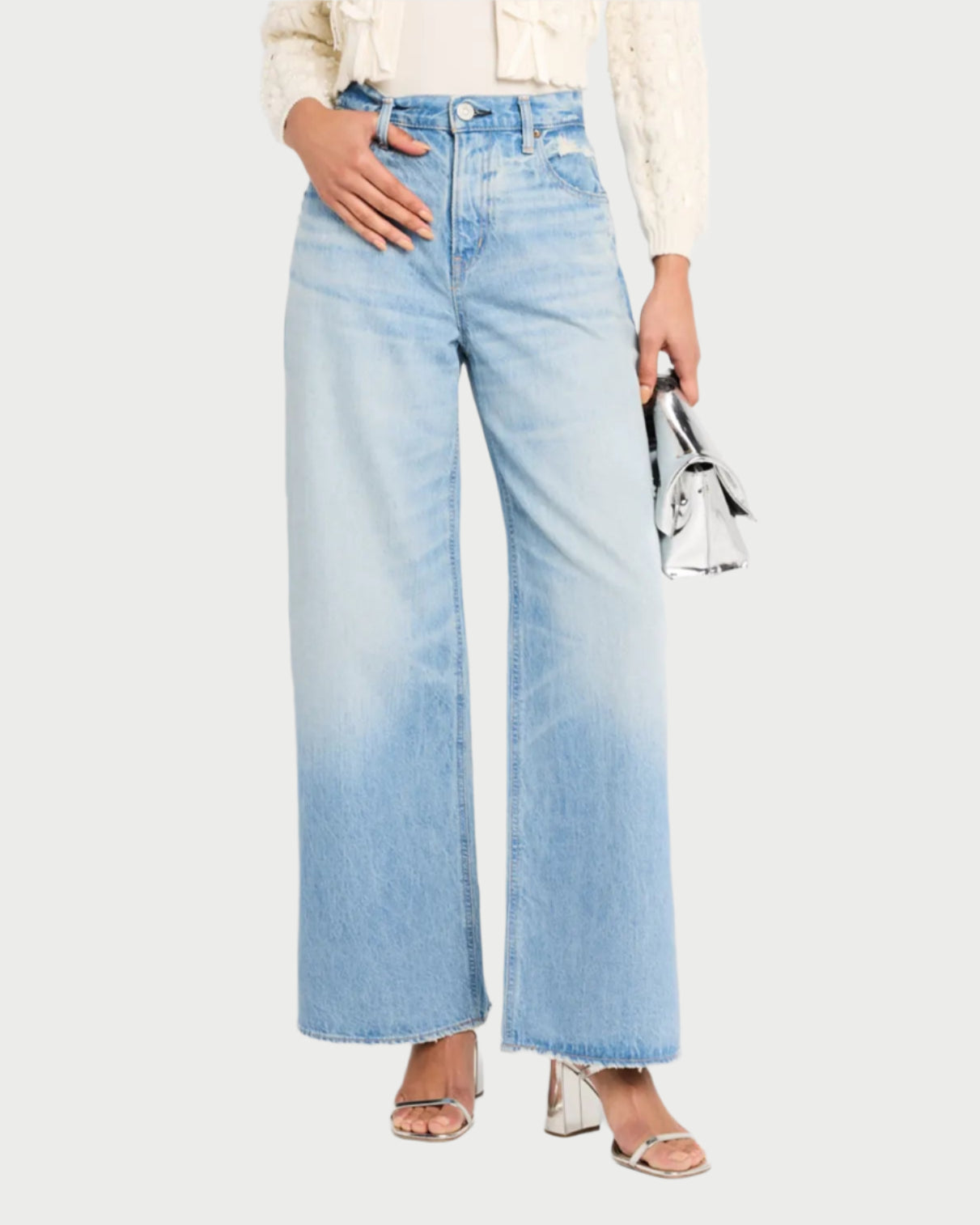 A person wears Moussy's Lowell Super Wide light blue jeans made from responsibly sourced cotton, a cream textured jacket, and silver high-heeled sandals, holding a small silver handbag against a light background.