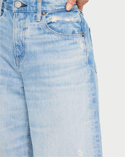 A close-up shows a person wearing Lowell Super Wide jeans by Moussy, featuring light blue denim with distressing and frayed patches. Made from responsibly sourced cotton, these jeans include button and rivet details, with a hand partially visible in the front pocket at the waistline.