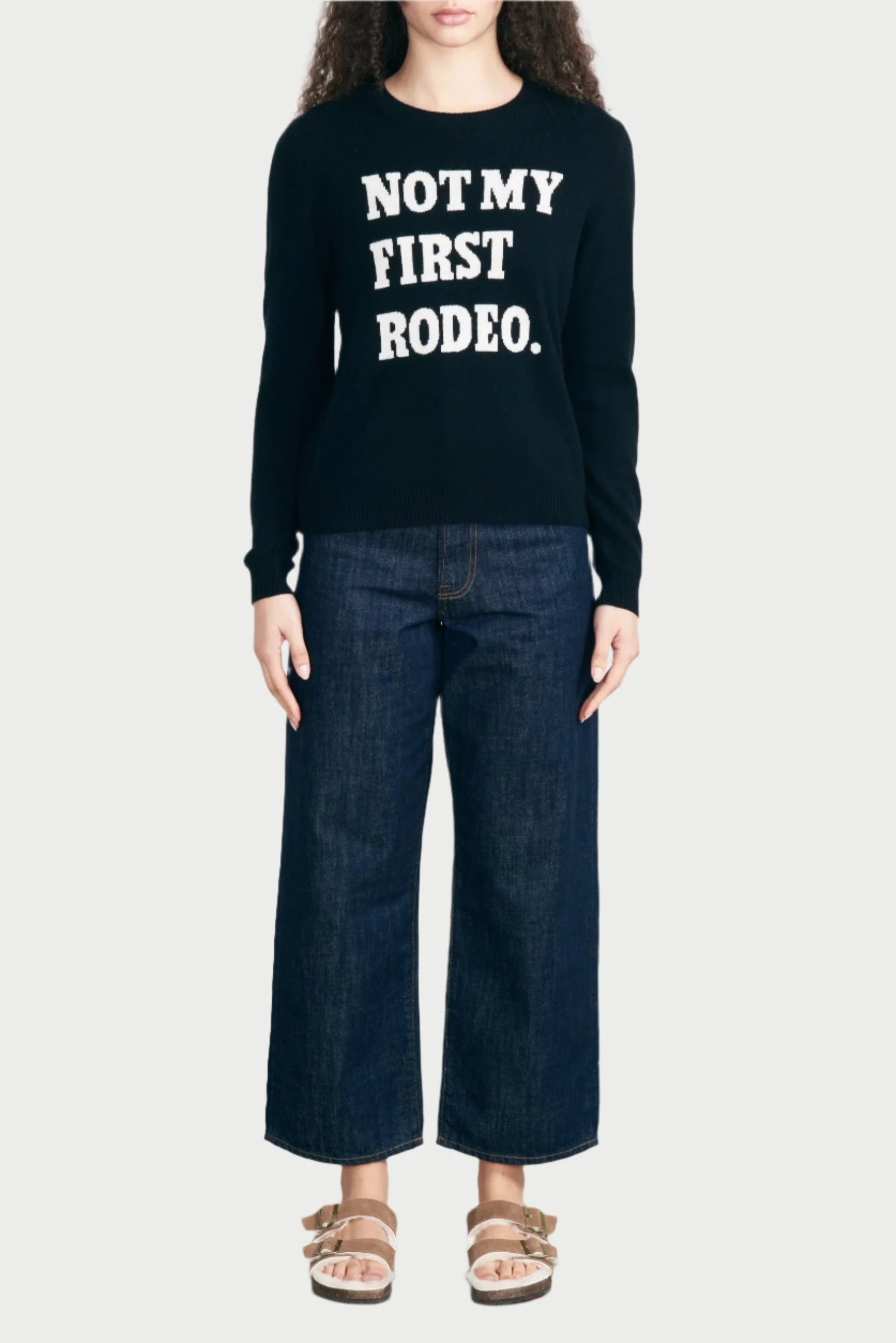 A person with long curly hair stands against a plain background, wearing the Rodeo Cashmere Crewneck by JUMPER, which has the text "NOT MY FIRST RODEO." They are also dressed in wide-legged blue jeans and beige sandals.