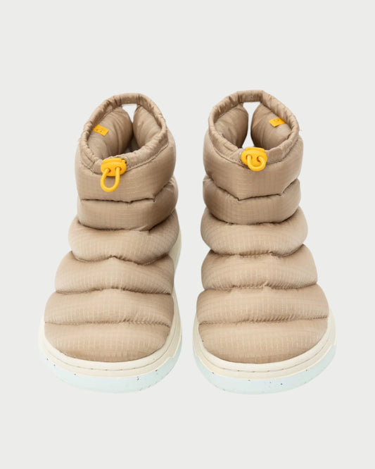 The Calgary Bootie by Oncept is a pair of beige, quilted, ankle-high slippers featuring white soles and yellow pull tabs on the front. With its padded, cushioned design tailored for indoor use, these shoes also include a molded footbed to ensure perfect support.