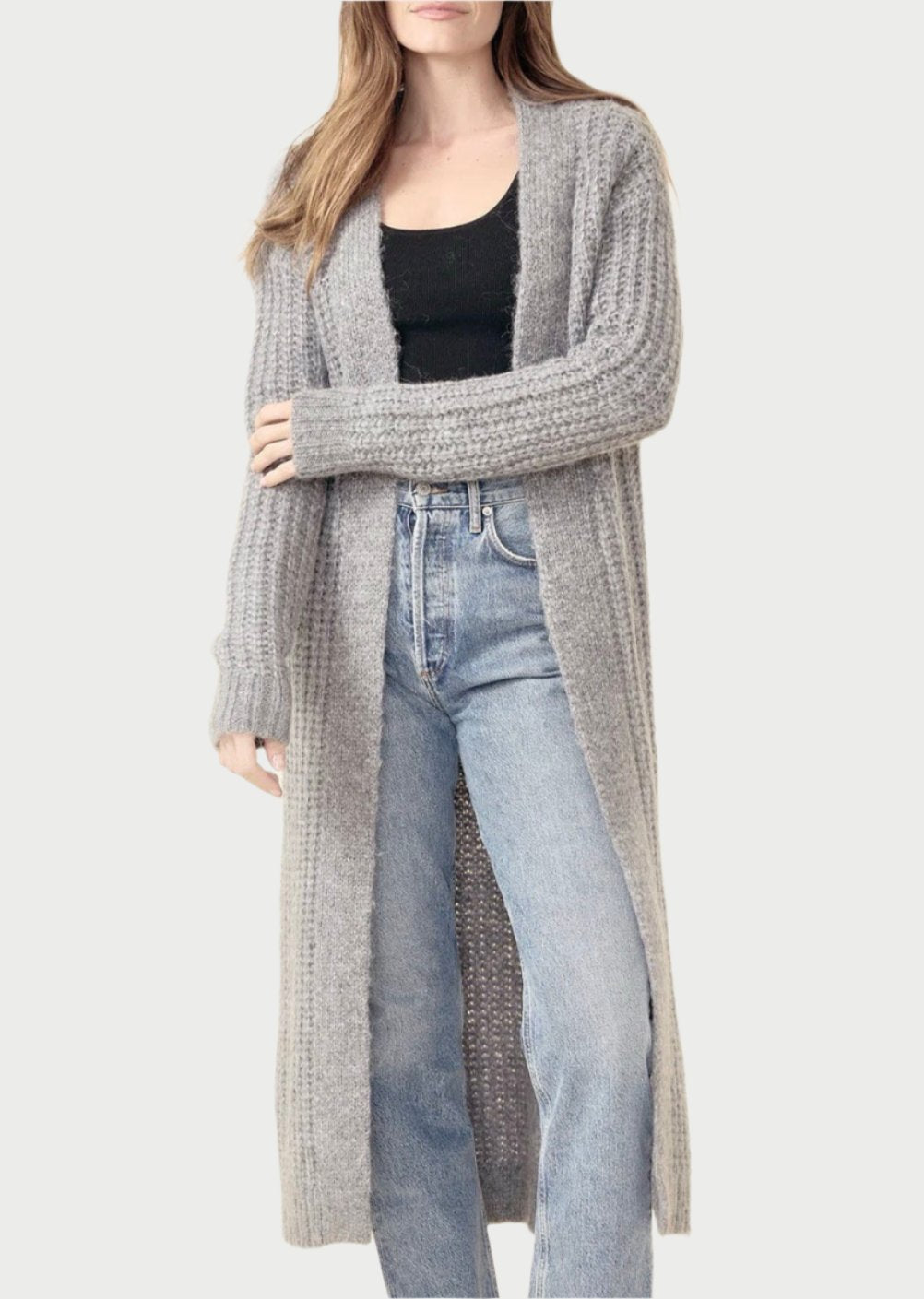 A person with long hair is standing and wearing a One Grey Day Chandler Duster over a black top and light blue high-waisted jeans. The cozy sophistication of the chunky, gray cardigan with open rib stitch detailing reaches below their knees as they exude a relaxed and confident expression against a plain, light background.