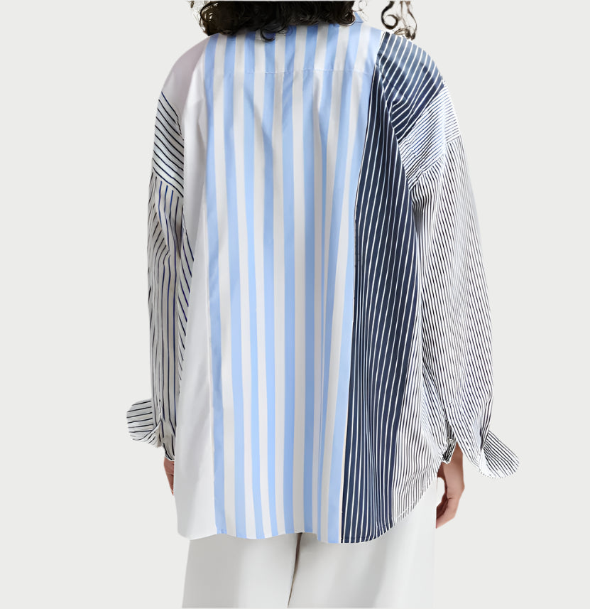 A person is wearing the Patchwork Shirt from Essentiel Antwerp, showcasing an oversized fit with mixed vertical stripes in blue, white, and black. Made from a lightweight cotton blend, they're standing with their back to the camera, paired effortlessly with white pants.